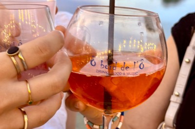 Deux personnes debout dans l'eau peu profonde d'un lac, tenant des verres, avec des arbres et un ciel bleu en arrière-plan.