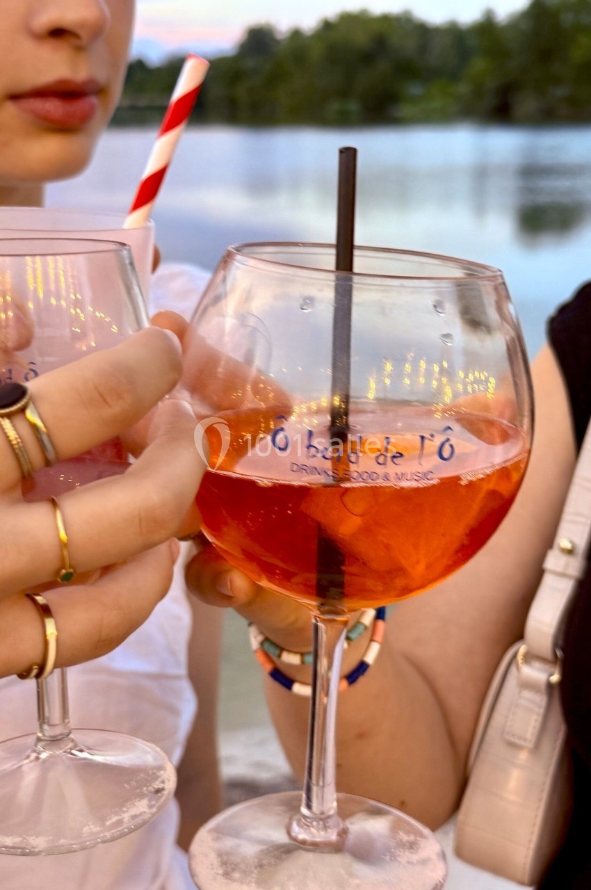 Deux personnes trinquent avec des verres de cocktail orange, près d'un plan d'eau au coucher du soleil.