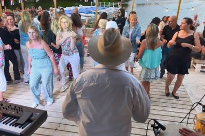 Deux personnes debout dans l'eau peu profonde d'un lac, tenant des verres, avec des arbres et un ciel bleu en arrière-plan.