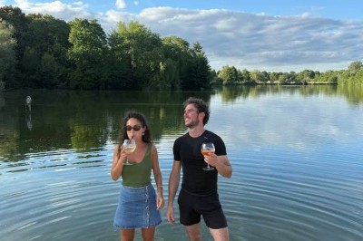 Deux personnes debout dans l'eau peu profonde d'un lac, tenant des verres, avec des arbres et un ciel bleu en arrière-plan.
