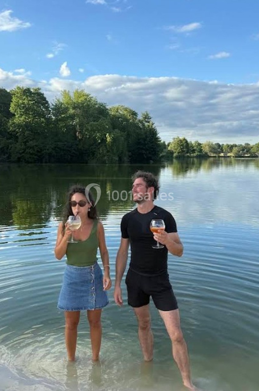 Deux personnes debout dans l'eau peu profonde d'un lac, tenant des verres, avec des arbres et un ciel bleu en arrière-plan.
