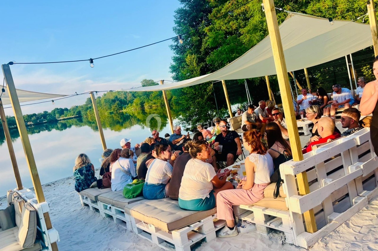 Groupe de personnes assises sur des banquettes en palettes sous des voiles d'ombrage, au bord d'un lac.