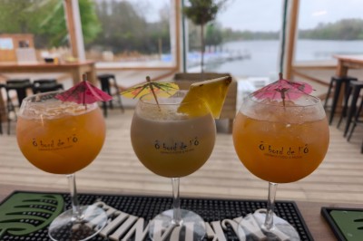 Terrasse d'un restaurant avec voiles d'ombrage, mobilier en bois, vue sur un étang et ambiance estivale.