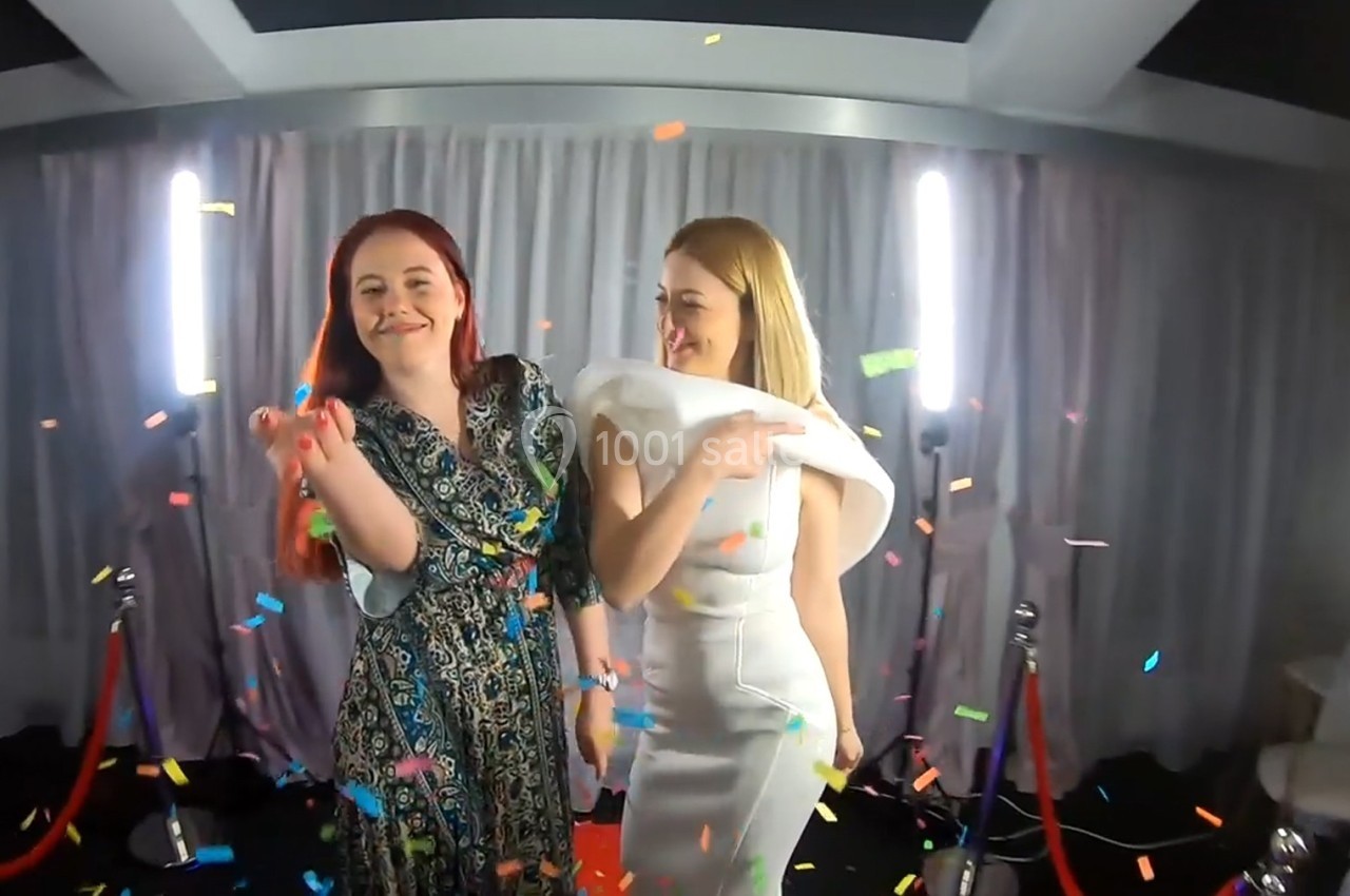 Deux femmes souriantes dansent sous des confettis colorés, devant un rideau gris et des cordons de sécurité rouges.