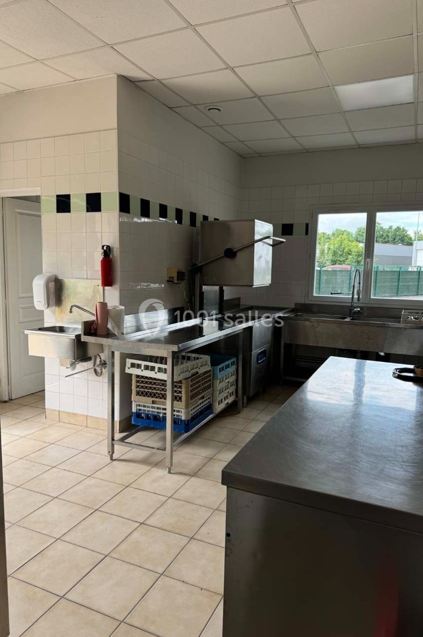 Cuisine professionnelle lumineuse avec évier, lave-vaisselle industriel, plans de travail en inox et carrelage clair.