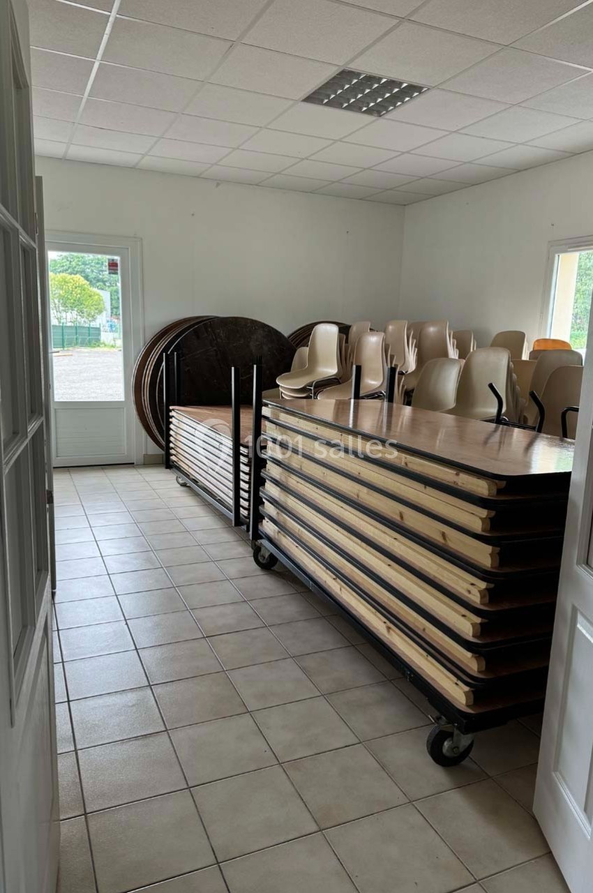 Tables empilées sur des chariots et chaises rangées dans une salle lumineuse avec carrelage au sol.