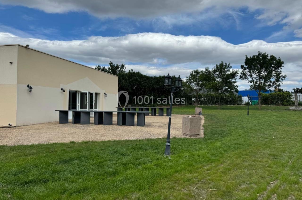 Vue d'une terrasse avec tables hautes, lampadaires et espace vert, entourée d'arbres sous un ciel nuageux.