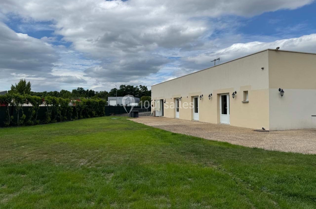 Maison moderne de plain-pied avec façade beige, entourée d'une pelouse et d'une haie, sous un ciel partiellement nuageux.