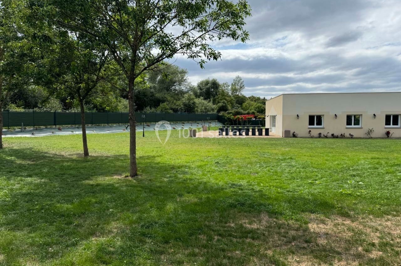 Espace extérieur avec pelouse, arbres, bâtiment clair et piscine clôturée en arrière-plan sous un ciel nuageux.