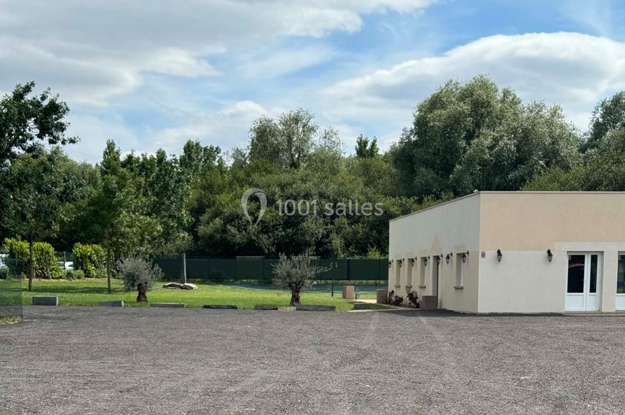 Bâtiment clair avec des murs beige, entouré d'arbres et d'une pelouse, donnant sur un grand parking en gravier.
