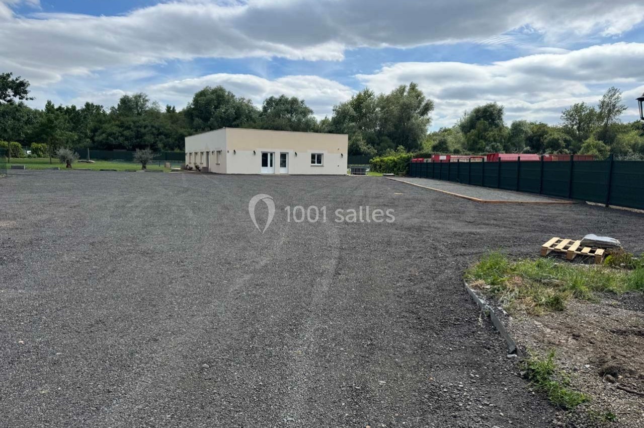 Terrain en gravier avec un bâtiment blanc au fond, entouré de végétation et clôturé par une barrière noire.