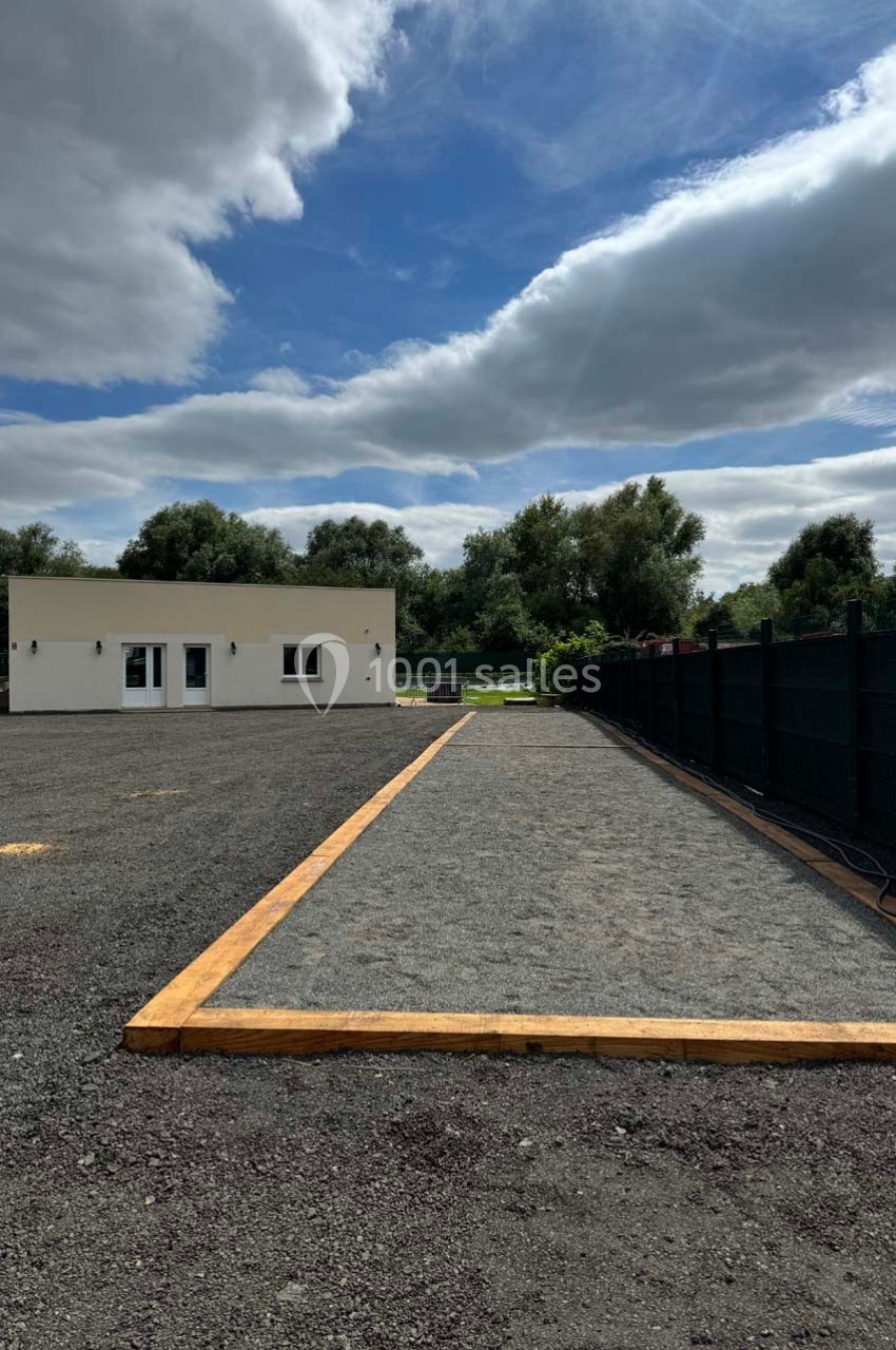 Terrain de pétanque en gravier bordé de bois, situé près d'un bâtiment blanc et entouré de végétation.