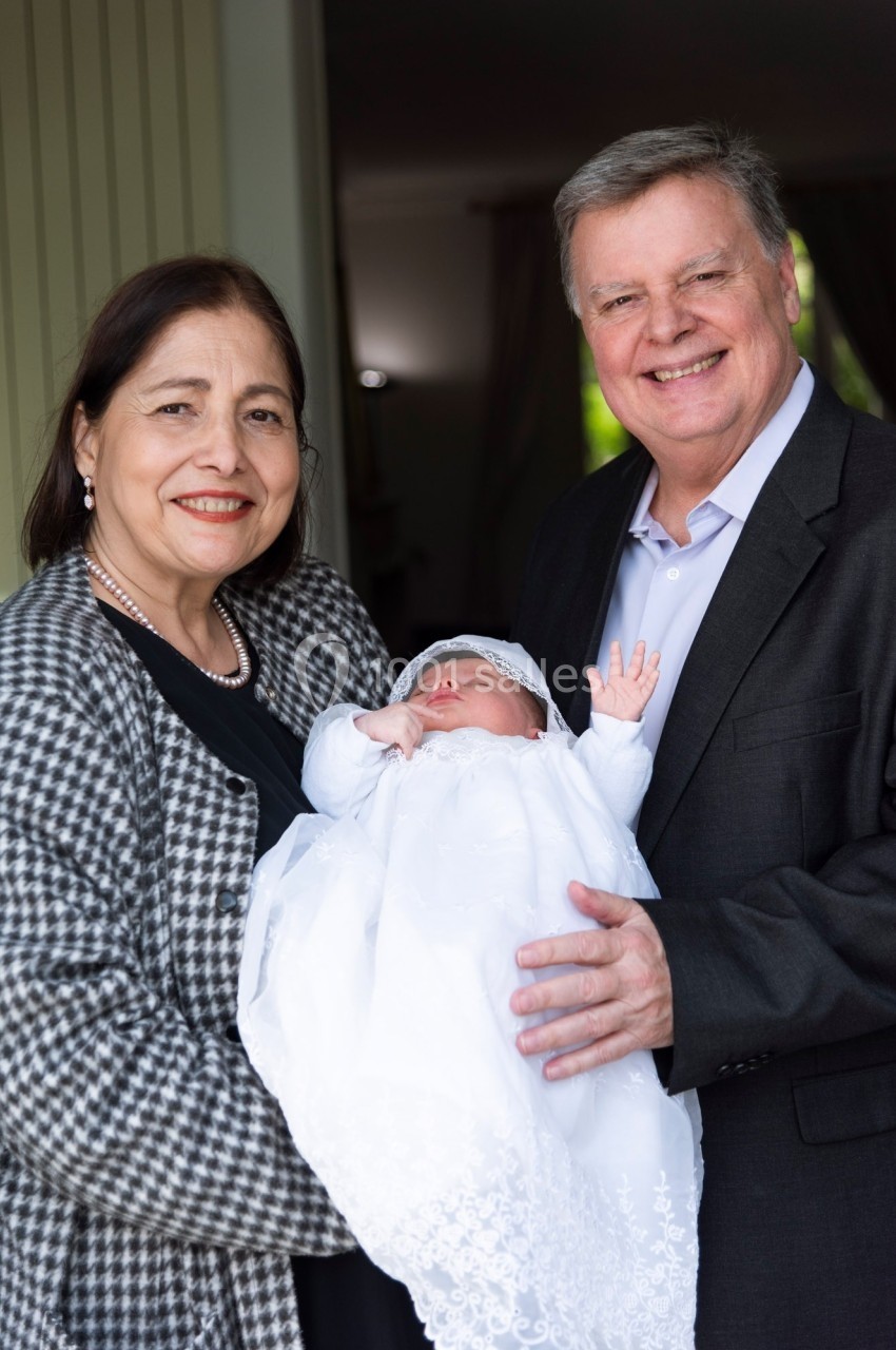 Un couple souriant tient un nourrisson vêtu d'une robe blanche dans un cadre intérieur lumineux.