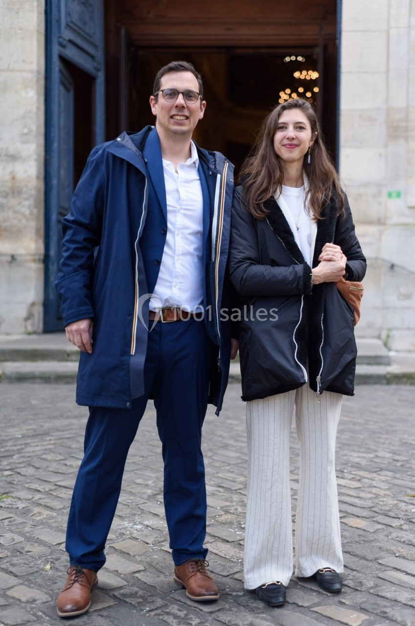Un homme et une femme debout devant une entrée de bâtiment en pierre, souriant et habillés chaudement.