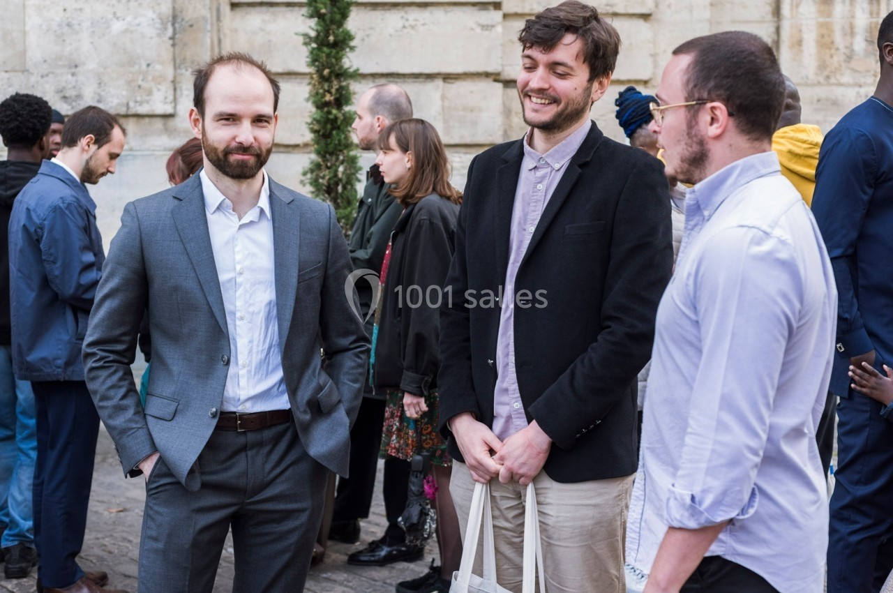 Trois hommes discutent ensemble en extérieur, entourés d'autres personnes dans un cadre urbain.