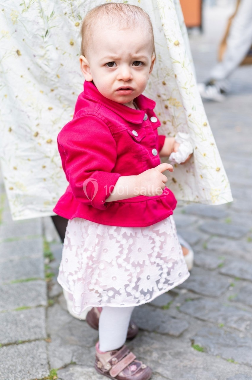 Un jeune enfant en robe rose et veste rouge regarde vers l'objectif, debout sur un sol pavé.