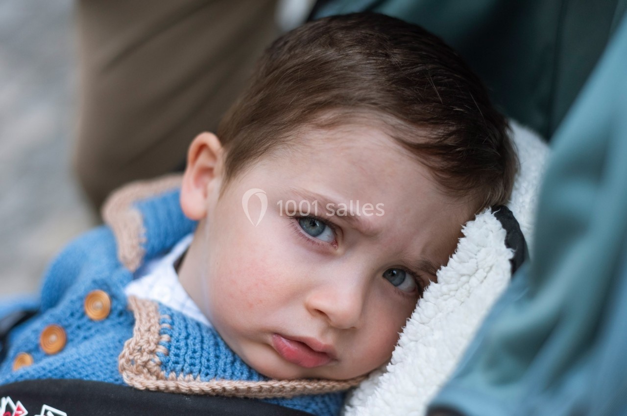 Enfant au regard sérieux, allongé sur un tissu blanc, vêtu d'un gilet bleu avec des boutons en bois.