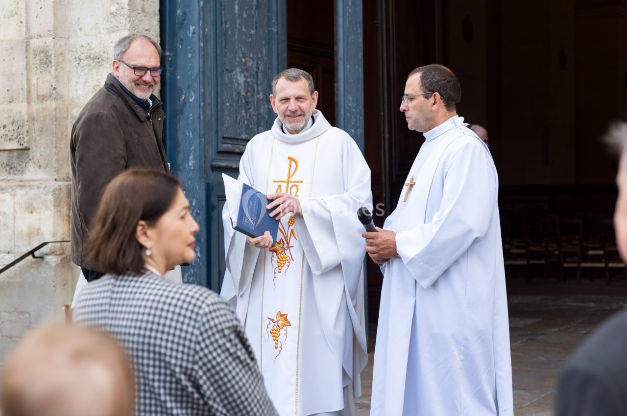 Un prêtre en tenue liturgique discute avec des personnes devant l'entrée ouverte d'une église.