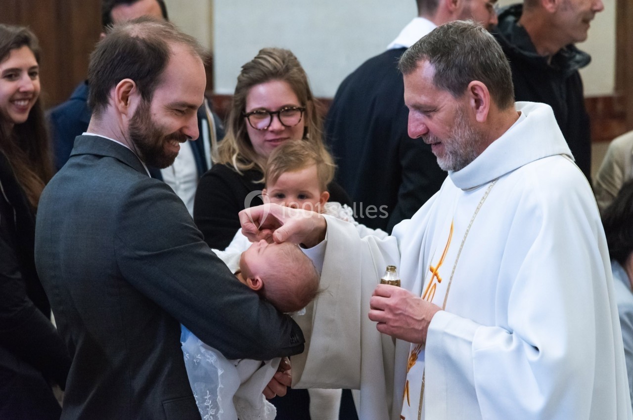 Un prêtre baptise un nourrisson dans les bras d'un homme, entouré de plusieurs personnes dans une église.