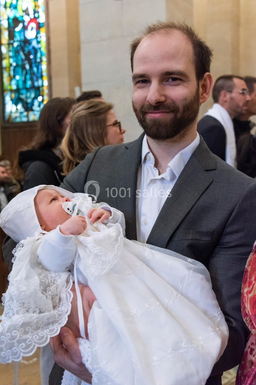 Un homme en costume tient un bébé en robe blanche de baptême dans une église avec des vitraux en arrière-plan.