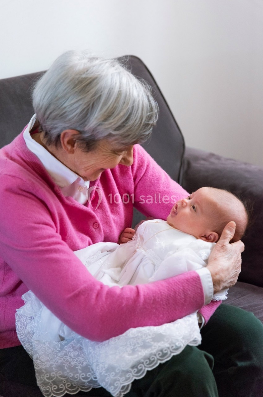 Une femme âgée aux cheveux gris tient un nourrisson vêtu de blanc dans ses bras, assise sur un canapé.