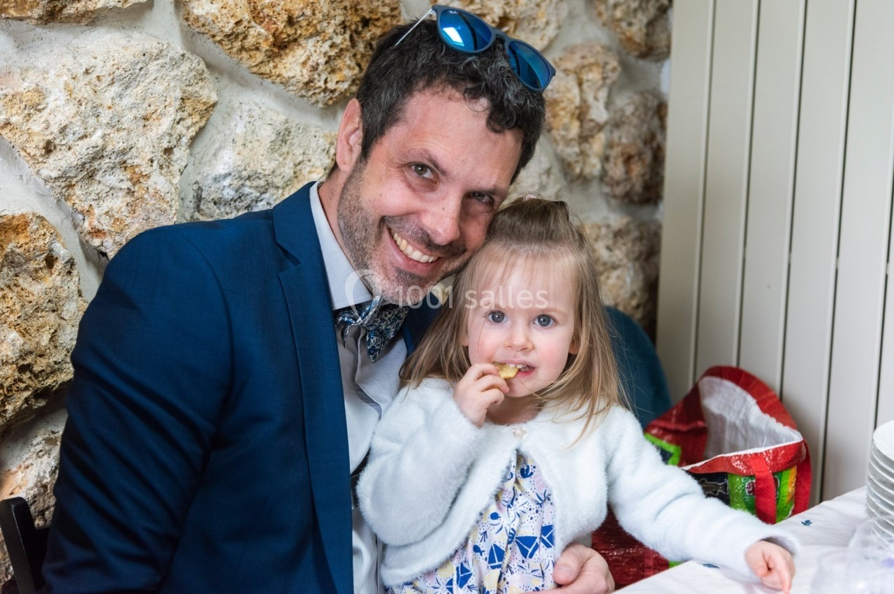 Un homme souriant en costume tient une petite fille mangeant un biscuit, assis devant un mur en pierre.