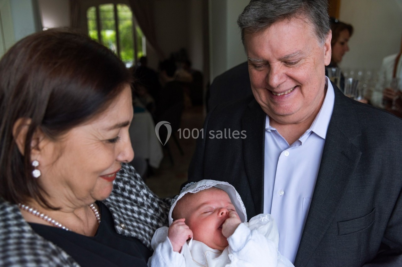 Un couple souriant tient un bébé vêtu de blanc lors d'un événement en intérieur.