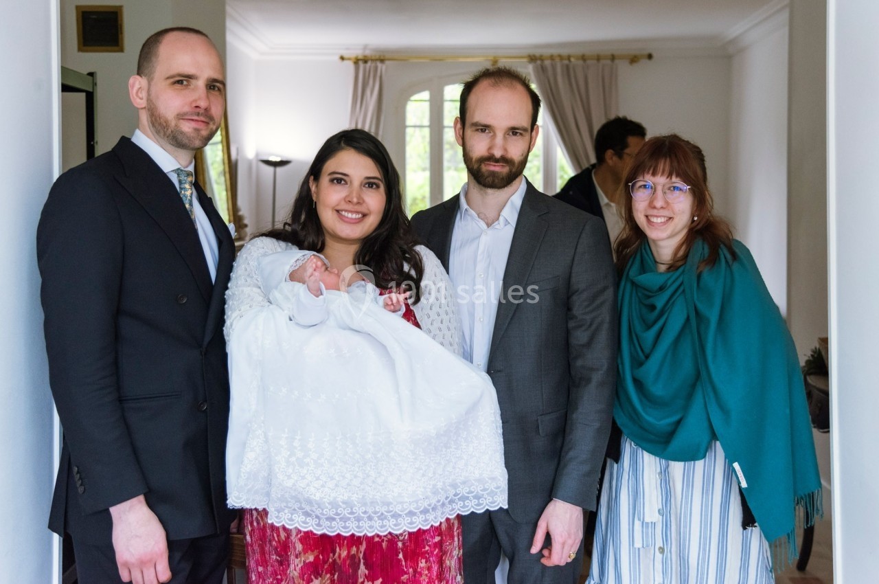 Quatre adultes posent avec un bébé en robe blanche dans une pièce lumineuse avec des rideaux ouverts en arrière-plan.
