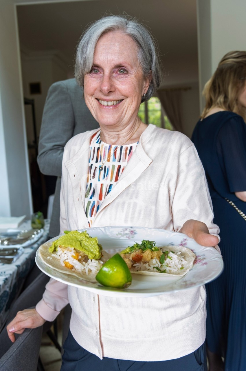 Une femme souriante tient une assiette contenant deux tacos garnis et un quartier de citron vert.