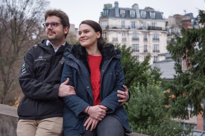 Un couple s'embrasse dans une rue pavée devant un bâtiment rose couvert de végétation.