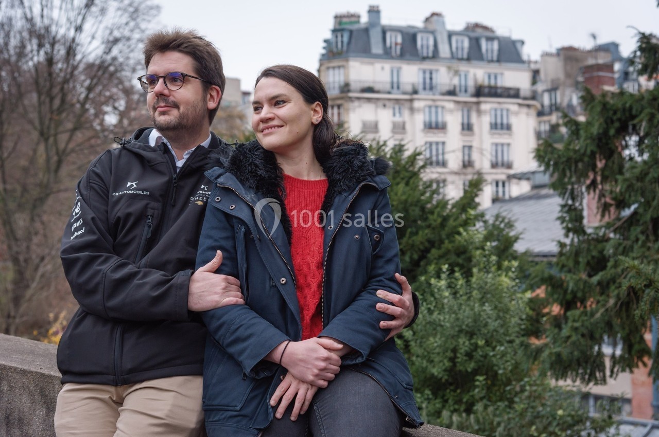 Un couple assis sur un muret dans un parc, avec des immeubles parisiens en arrière-plan.
