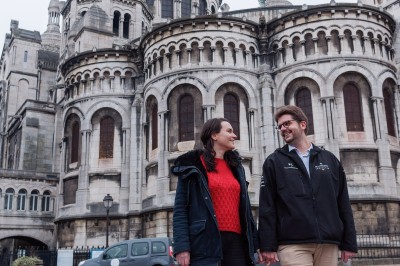 Un couple s'embrasse dans une rue pavée devant un bâtiment rose couvert de végétation.