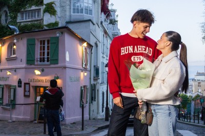 Un couple s'embrasse dans une rue pavée devant un bâtiment rose couvert de végétation.