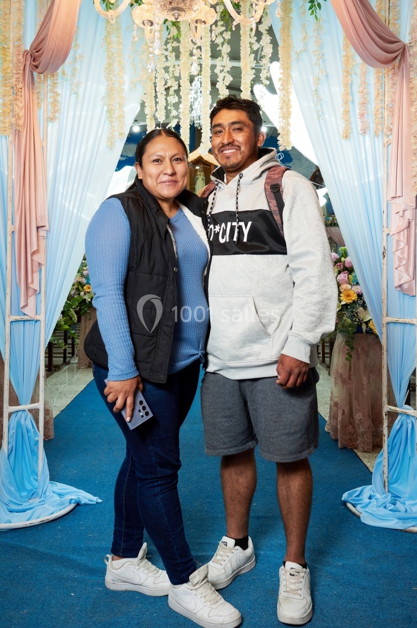 Un homme et une femme souriants posent sous une arche décorée de fleurs et de tissus colorés.