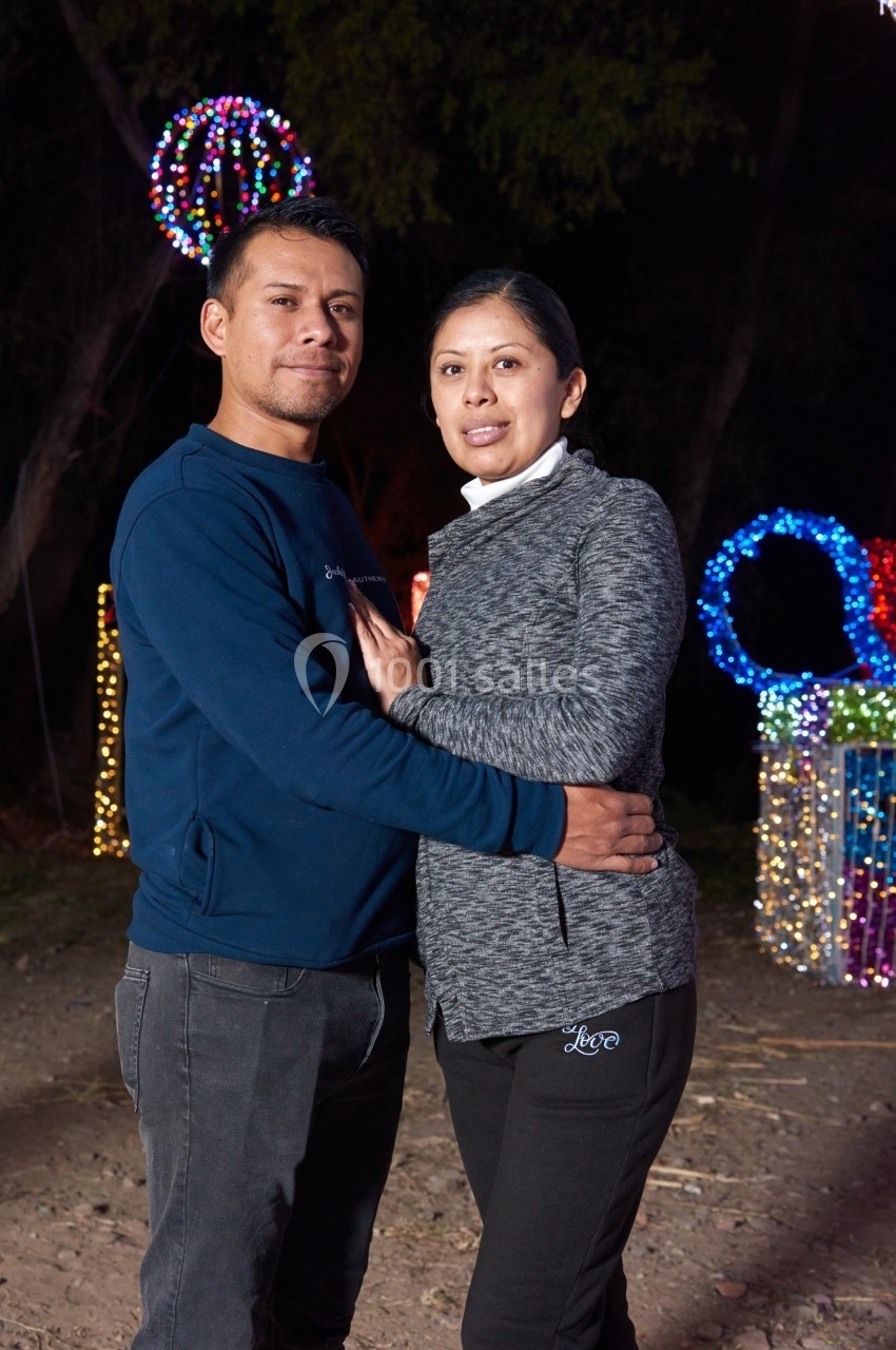 Un couple pose debout en extérieur, entouré de décorations lumineuses colorées dans un cadre nocturne.