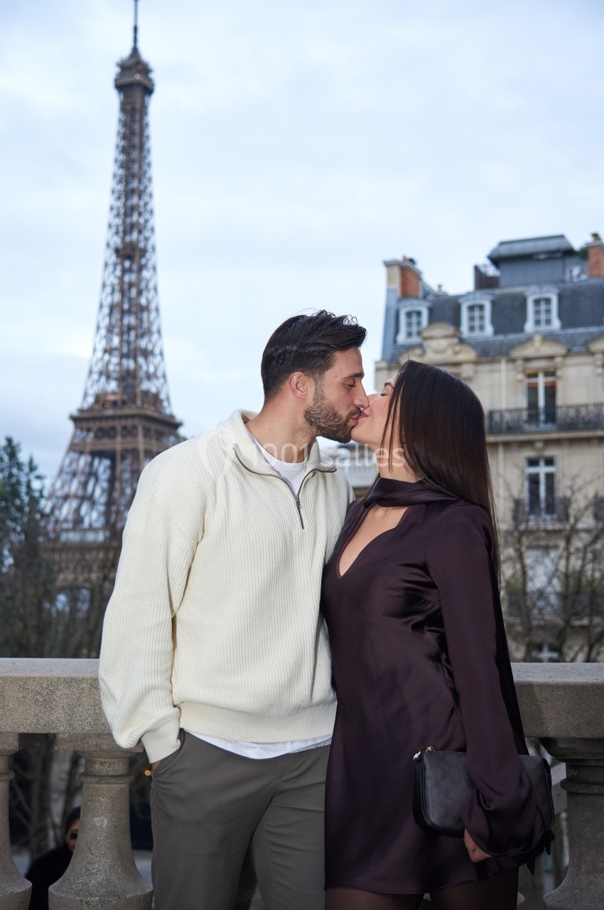Un couple s'embrasse sur une terrasse avec la tour Eiffel et des immeubles parisiens en arrière-plan.
