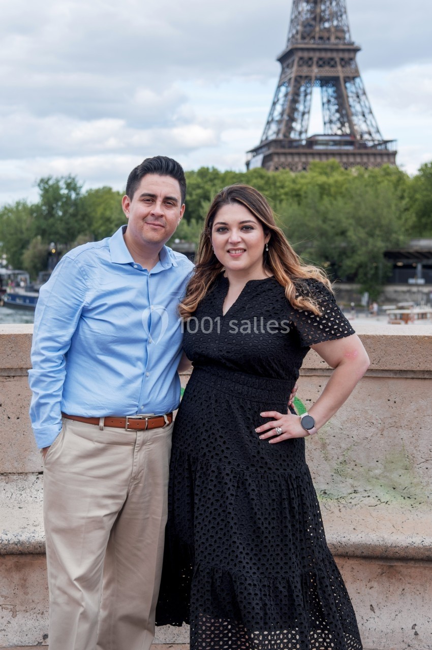 Un couple pose devant la tour Eiffel, avec des arbres et un ciel nuageux en arrière-plan.