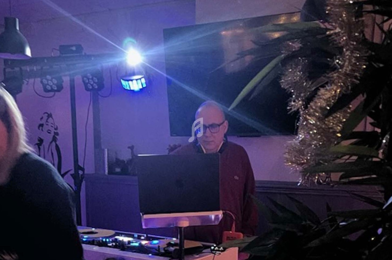 Un homme se tient derrière une table de mixage avec un ordinateur portable, dans une pièce faiblement éclairée.