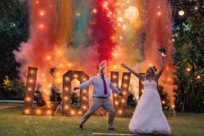 Un couple en tenue de mariage pose devant des lettres lumineuses ’LOVE’ et des feux colorés dans un jardin.