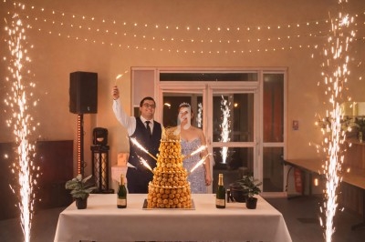 Un couple pose devant une pièce montée éclairée par des cierges magiques dans une salle décorée de guirlandes lumineuses.