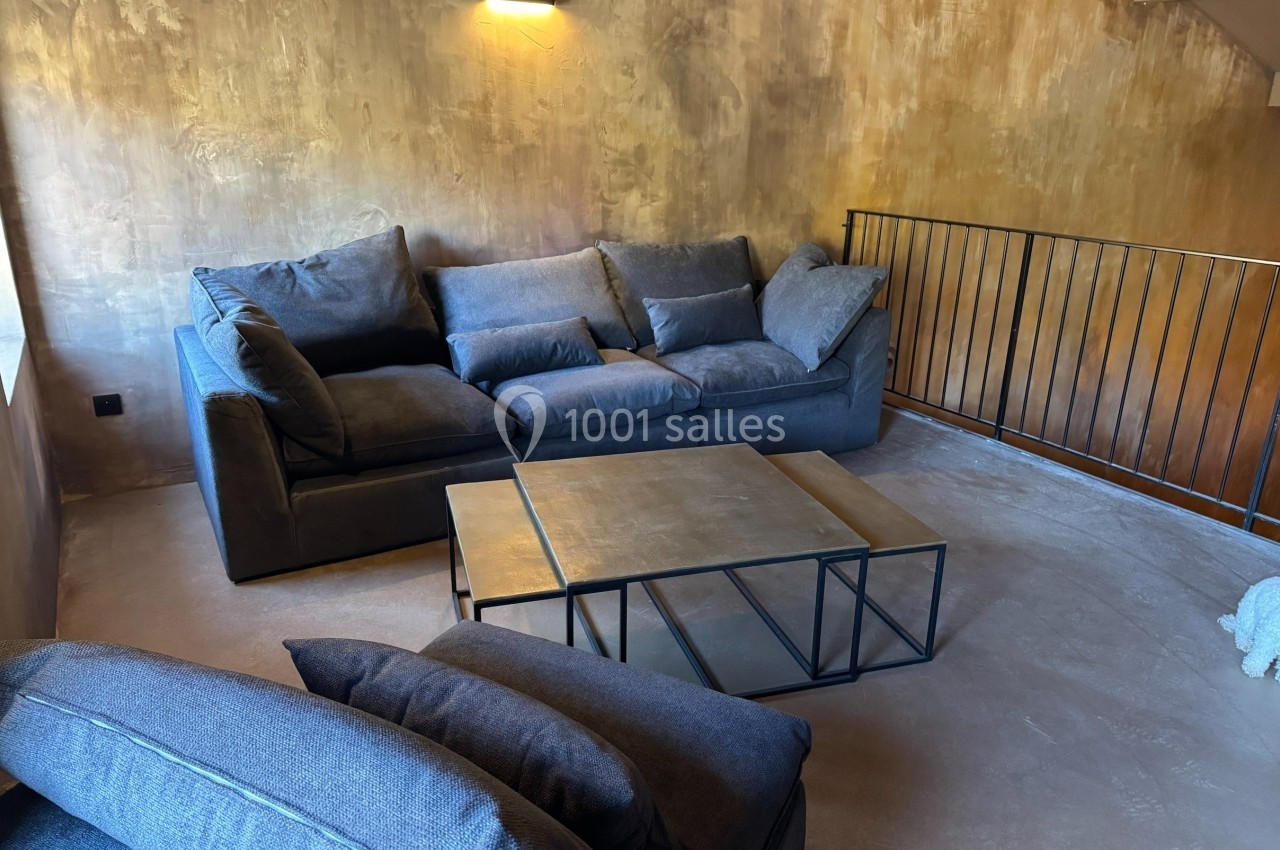 Salon avec canapé gris, table basse en métal et mur texturé beige, éclairé par une applique murale.