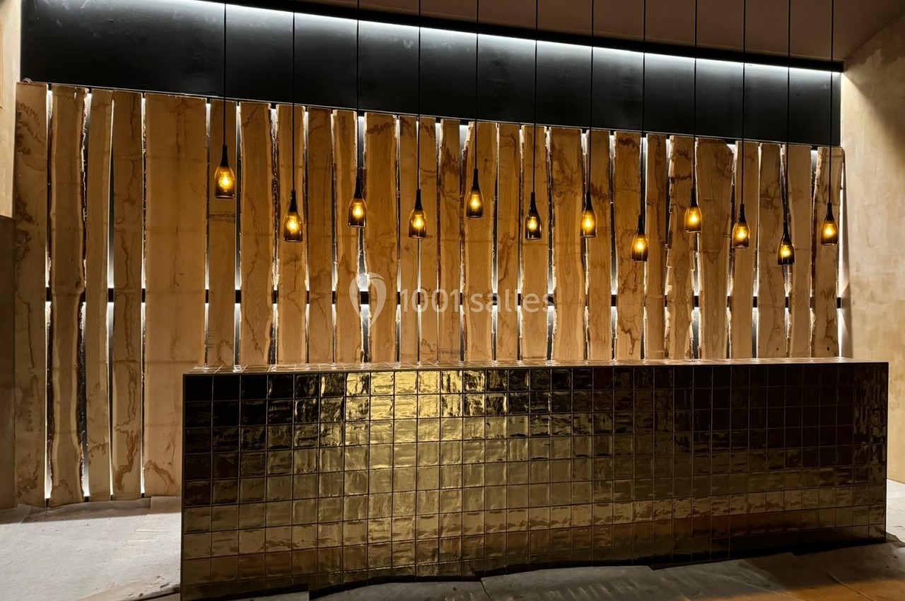 Comptoir recouvert de carreaux dorés devant un mur décoratif en bois avec des suspensions lumineuses alignées.