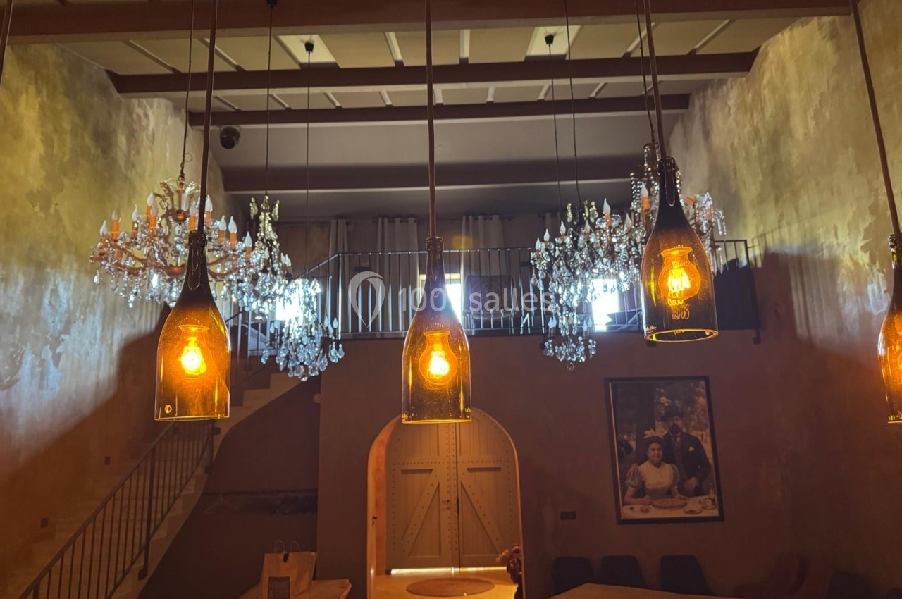 Suspensions lumineuses en bouteilles et lustres en cristal éclairant une salle avec murs texturés et mezzanine.