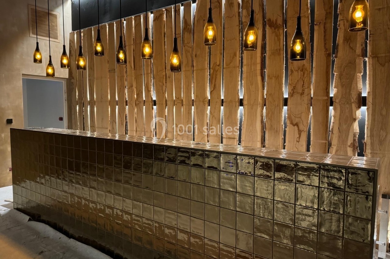 Comptoir recouvert de carreaux dorés, éclairé par des suspensions avec ampoules, devant un mur en bois ajouré.