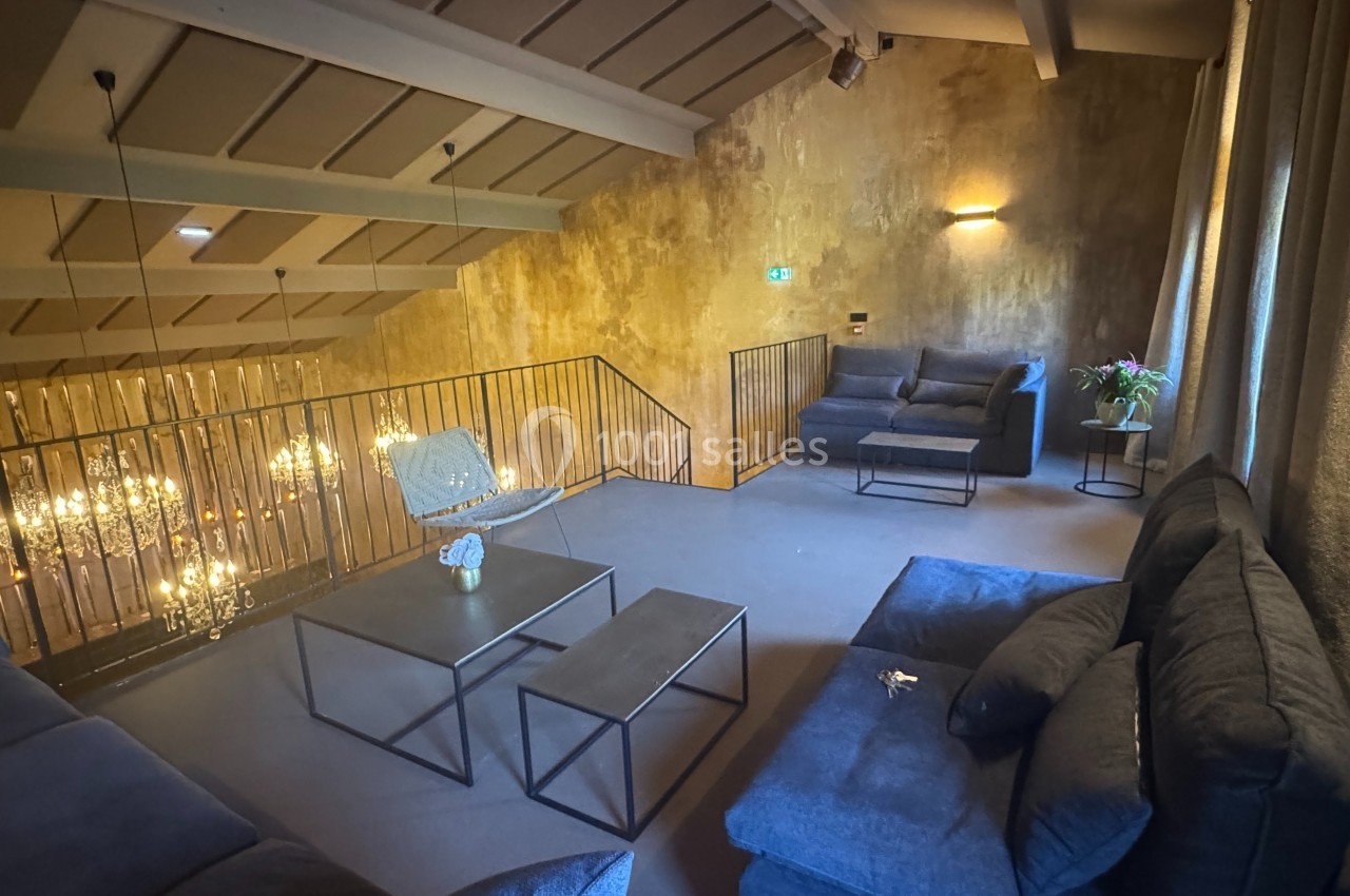 Espace salon en mezzanine avec canapés gris, tables basses noires, mur texturé et lustres suspendus visibles en contrebas.