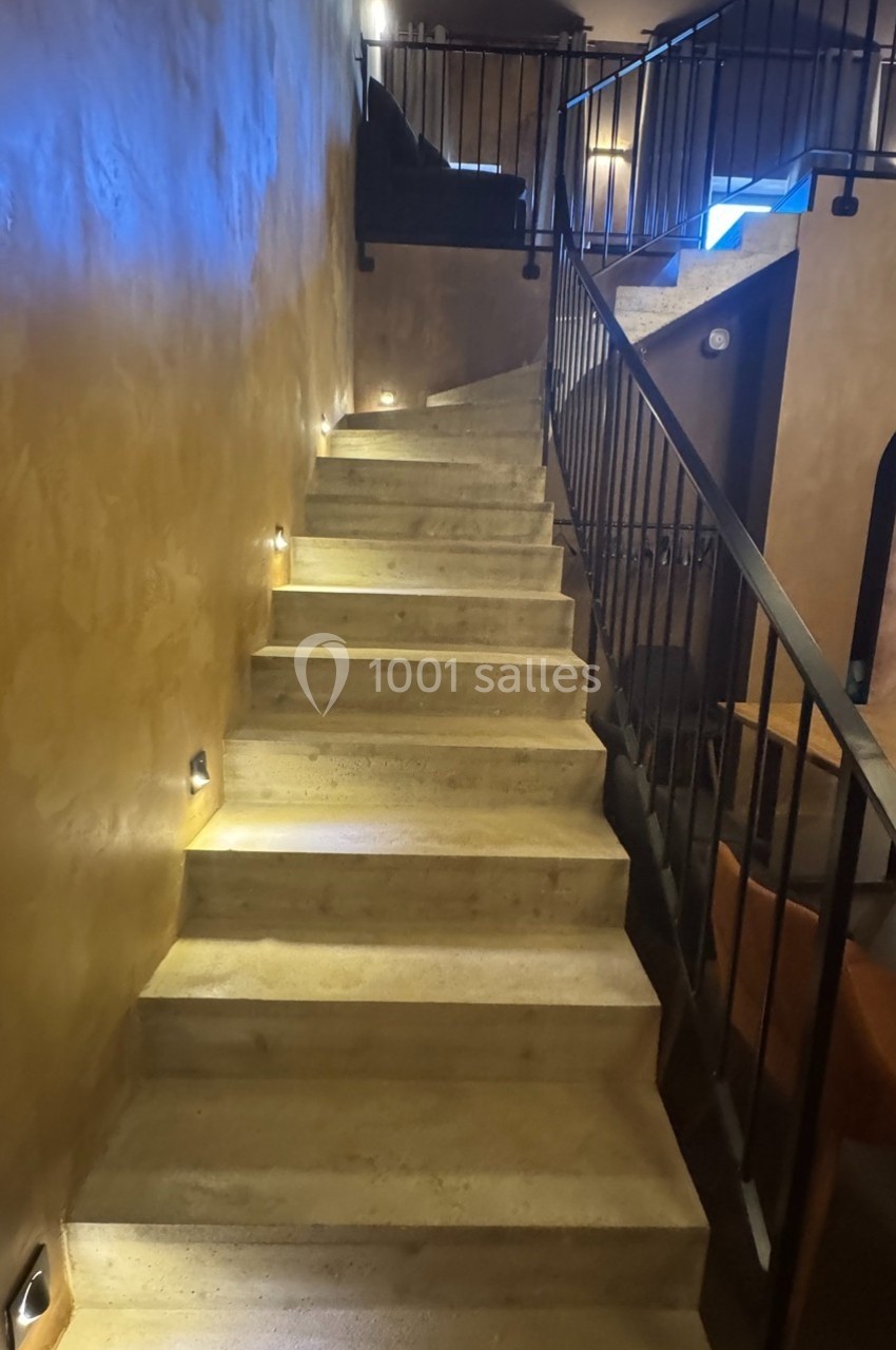 Escalier en béton éclairé par des lumières murales, avec une rampe en métal et un décor intérieur aux tons chauds.