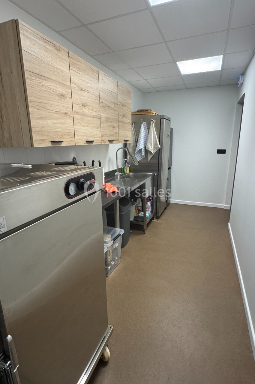 Cuisine professionnelle étroite avec plan de travail, évier, réfrigérateur, placards en bois et sol en lino marron.