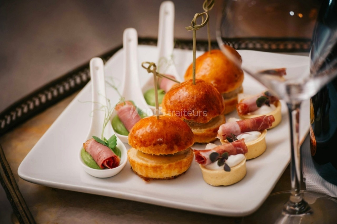 Assortiment d'amuse-bouches comprenant mini-burgers, cuillères garnies et tartelettes sur une assiette blanche.