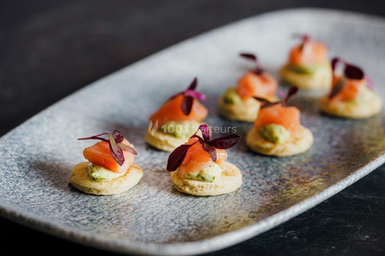 Assortiment de canapés au saumon fumé, avocat et herbes sur un plateau en céramique gris.
