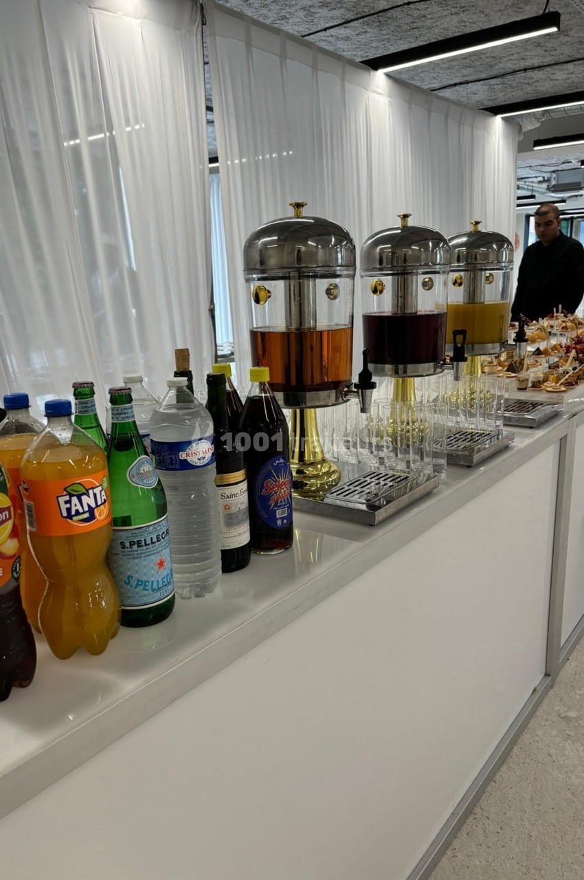 Buffet avec distributeurs de boissons, bouteilles variées et verres alignés sur une table blanche dans un espace lumineux.